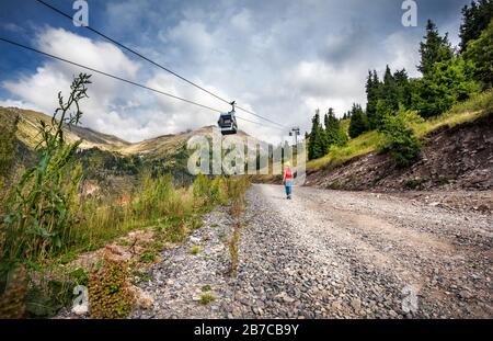 Femme de tourisme avec sac à dos et chapeau jaune marchant sur la route près du téléphérique dans la vallée de la montagne Banque D'Images