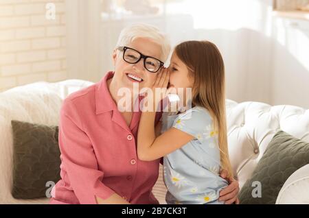 Jolie fille chuchotant quelque chose à l'oreille de sa grand-mère dans le salon Banque D'Images