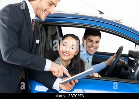 Un couple heureux achète une voiture en parlant avec un concessionnaire assis en auto Banque D'Images