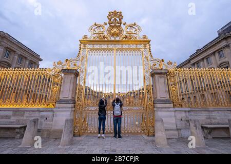 Deux touristes prenant des photos de téléphones cellulaires à travers les bars dorés de la porte du château de Versailles dans la périphérie de Paris, France, Europe Banque D'Images