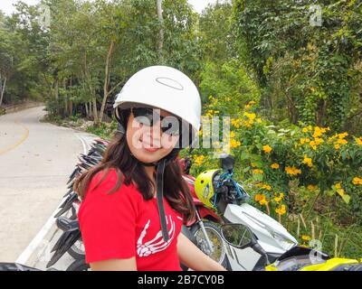 Jeune femme thaïlandaise portant un casque. Derrière elle, une moto verte et un fond tropical Banque D'Images