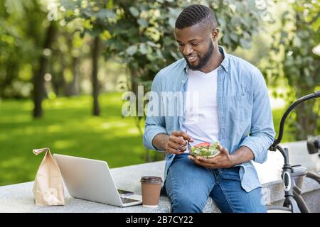 Black Guy manger salade et boire du café à l'extérieur, se détendre avec ordinateur portable Banque D'Images