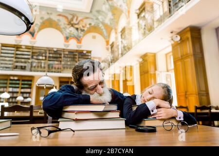 Vieux professeur d'homme barbu senior et son étudiant ou petite-fille dorment dans une bibliothèque en liant sur la table avec beaucoup de livres dans l'ancien v Banque D'Images