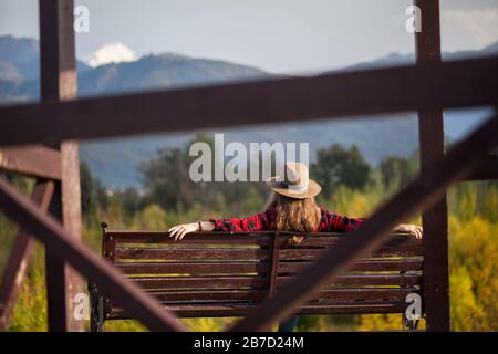 Femme en chemise à carreaux rouges et un chapeau assis sur le banc en bois au fond des montagnes et des forêts d'automne Banque D'Images