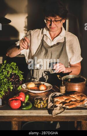 Femme sureau en lin tablier cuisine, dégustation, servant la soupe traditionnelle turque à la viande Haslama avec des légumes dans la cuisine moderne intérieur. Confort de chute f Banque D'Images