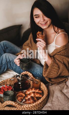 Jeune femme jeune brunette souriante dans des vêtements confortables et décontractés, prenant le petit déjeuner au lit avec thé, croissants, confiture et fraises mûres fraîches Banque D'Images