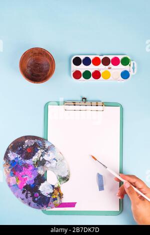 Set de peinture artisanal. La main de la femme avec un pinceau au-dessus des peintures aquarelles, une palette d'artistes et une feuille de papier vierge sur fond bleu clair, Banque D'Images