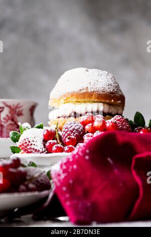 Gâteau éponge Victoria avec confiture de framboises et crème fouettée, généreusement arrosé de sucre en poudre , servi avec des fruits. Banque D'Images