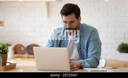 Homme sérieux portant des lunettes de travail sur ordinateur portable en ligne dans la cuisine Banque D'Images
