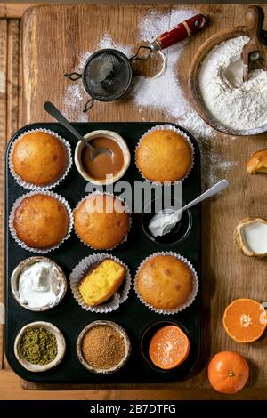 Oranges ou clémentines faites maison muffins gâteaux dans un plateau cuit avec farine, ingrédients et différentes garnitures au-dessus de planche à découper en bois Banque D'Images