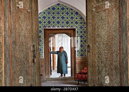 Femme ouzbèke vêtue de vêtements locaux et regardant par les portes en bois du mausolée Madari Kahn, à Kokand, en Ouzbékistan. Banque D'Images