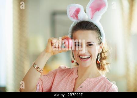 femme de ménage souriante de âge moyen dans un chemisier rose et des oreilles de lapin de pâques dans le salon moderne en journée de printemps ensoleillée montrant l'oeuf de pâques rouge. Banque D'Images