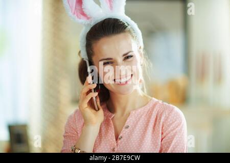 Portrait d'une femme moderne souriante dans un chemisier rose et des oreilles de lapin de pâques à la maison moderne en journée de printemps ensoleillé à l'aide d'un téléphone cellulaire. Banque D'Images