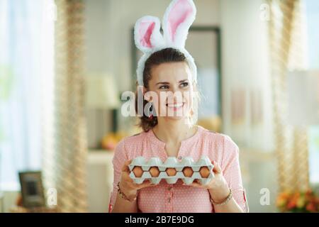 pensive élégante femme dans un chemisier rose et des oreilles de lapin de pâques à la maison moderne dans le soleil de printemps tenant la grande boîte de carton d'œufs. Banque D'Images