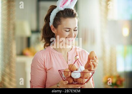 femme de ménage élégante souriante dans un chemisier rose et des oreilles de lapin de pâques tenant le panier avec des œufs et regardant l'oeuf dans la maison moderne en journée de printemps ensoleillée. Banque D'Images