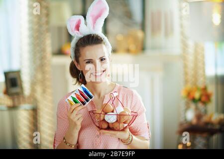 joyeux jeune femme de ménage dans un chemisier rose et des oreilles de lapin de pâques montrant le panier avec des œufs et des colorants alimentaires dans le salon moderne en journée de printemps ensoleillée. Banque D'Images