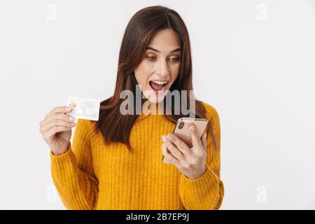 Image d'une femme adulte avec brunette excitée portant un pull tenant le téléphone portable et la carte de crédit isolée sur fond blanc Banque D'Images