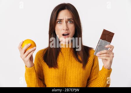 Image d'une femme adulte douteuse de brunette hésitant tout en tenant une barre d'orange et de chocolat isolée sur fond blanc Banque D'Images