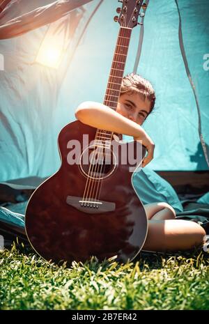 Une jeune fille gentille assise sur l'herbe verte fraîche avec de la guitare, un enfant talentueux joue un instrument de musique, profitant du temps actif au camp d'été Banque D'Images