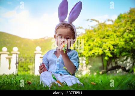 Mignon petit garçon de bébé vêtu de grandes oreilles de lapin, joyeux enfant mangeant des carottes et célébrant les vacances chrétiennes, costume traditionnel de Pâques, le petit enfant aime Banque D'Images