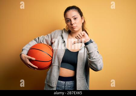 Jeune sportif brunette qui tient le ballon de basket-ball sur fond jaune avec le visage en colère, signe négatif montrant déplaît avec les pouces vers le bas, r Banque D'Images