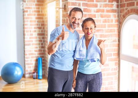 Couple sportif d'âge moyen debout avant de faire de l'exercice à la salle de gym faire des pouces heureux vers le haut geste avec la main. Approbation de l'expression en regardant la caméra avec s Banque D'Images