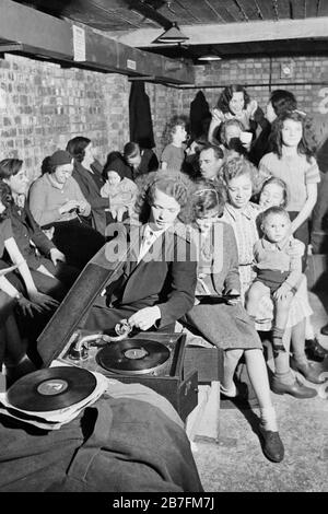 Une jeune femme joue un gramophone dans un abri anti-aérien dans le nord de Londres au cours de 1940. Une jeune femme met l'aiguille gramophone sur un enregistrement pour apporter un peu de lumière de secours un abri anti-aérien, quelque part dans le nord de Londres. Le reste de l'hébergeurs semblent profiter de son choix de musique. Dans l'arrière-plan, une femme peut être vu le tricot, comme d'autres chat pour faire passer le temps. Banque D'Images