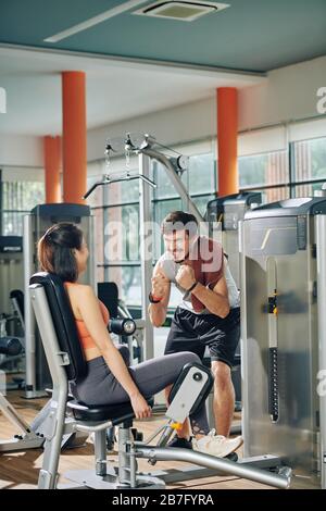 L'entraîneur de forme physique émotive excité motivant et encourageant la jeune femme à faire de l'exercice dans l'intérieur et l'extérieur de la machine de cuisse Banque D'Images