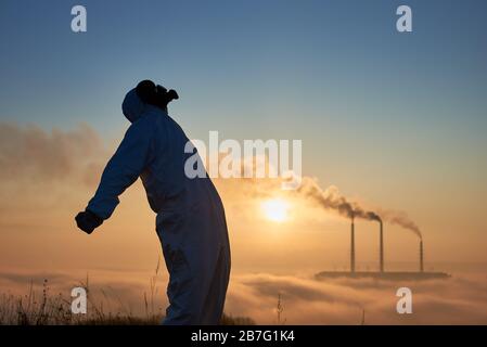 Vue latérale du scientifique dans le masque à gaz. Ciel bleu au-dessus de la centrale électrique en arrière-plan. Écologiste masculin dans un costume de protection se propageant les mains aux côtés dans le désespoir tandis que les cheminées produisant de la fumée dans l'atmosphère Banque D'Images