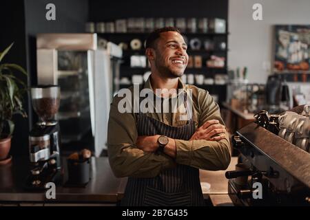 Jeune propriétaire afro-américain de petit café debout derrière le comptoir portant un tablier avec des bras croisés regardant loin Banque D'Images