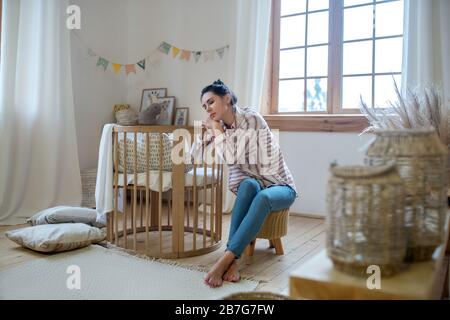 Jeune maman fatiguée assise au lit avec des yeux fermés, reposant sa tête sur les mains Banque D'Images