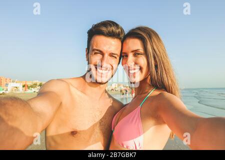 Un couple de voyage heureux qui fait du selfie avec la plage en arrière-plan - concept de couleurs ensoleillées d'été et d'humeur romantique - les jeunes riant et faisant de l'em Banque D'Images