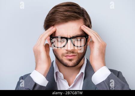 Portrait rapproché d'un jeune homme triste en costume avec des poils et des lunettes touchent sa tête et se sentent forte migraine tout en se tenant sur fond gris Banque D'Images