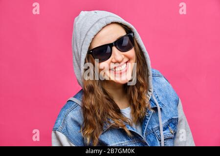 Fille caucasienne élégante avec un Jean bleu et des lunettes 8 bits et sourire à l'appareil photo sur un fond rose. Gros plan. Banque D'Images