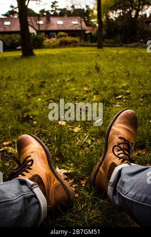 Se rafraîchir dans le jardin avec vue sur la maison et quelques arbres, vue de première personne sur les jambes et les bottes, chaussures en cuir, pantalon roulé Banque D'Images