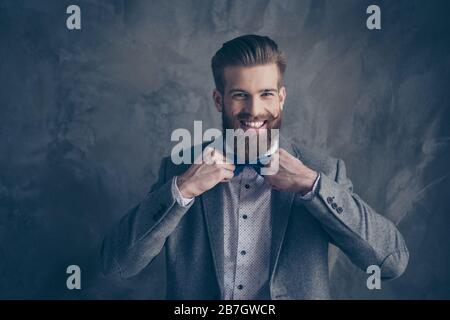 Heureux jeune homme heureux barbu avec moustache en forme de formalewear se tient sur un fond gris et tient son noeud-cravate avec les deux mains Banque D'Images