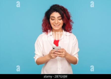 Portrait d'une belle femme hipster avec des cheveux rouges fantaisie à l'aide d'un smartphone, lire ou taper un message drôle, discuter en réseau social avec un look heureux. Banque D'Images