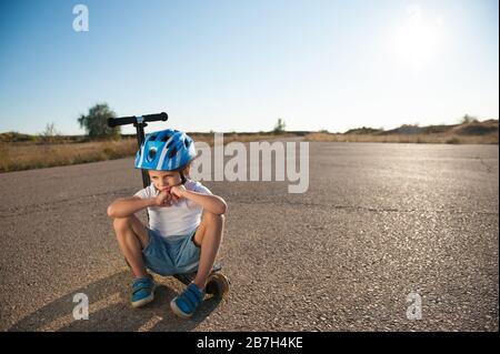 un enfant triste et peu actif dans un casque de protection sportif assis seul sur un scooter sur une route asphaltée désertique d'été vide au coucher du soleil Banque D'Images