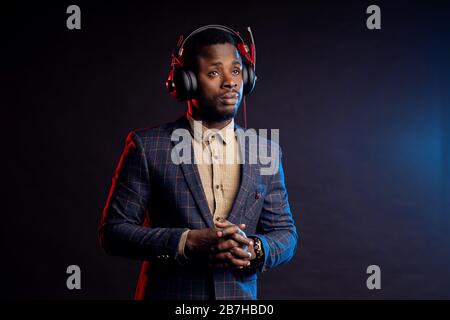 Gros plan sur un jeune homme afro-américain non rasé écoutant de la musique dans un casque, regardant vers le bas, se posant dans le noir. Portrait de DJ élégant avec casque, Banque D'Images