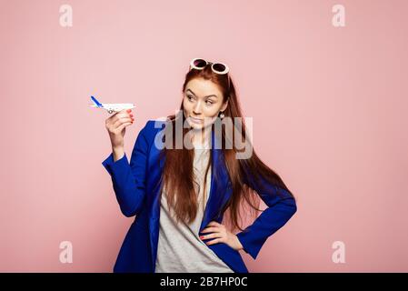 Jeune fille gaie à tête rouge tenant l'avion et posant isolé sur fond rose. Une femme surprise regarde un avion modèle et demande où mes bagages sont. Banque D'Images