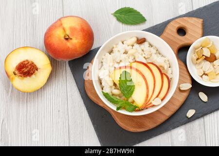 Porridge de sorgho avec morceaux de pêche, noix de cajou et amandes dans des bols en porcelaine, pêches fraîches sur bois et panneaux de pierre. Sorgho végétalien sans gluten Banque D'Images