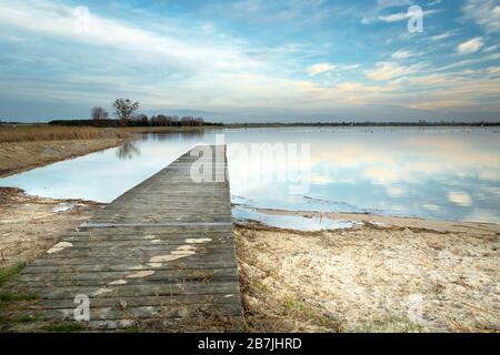 Pont en bois surgrandi avec de l'herbe vers le lac calme et nuages sur le ciel Banque D'Images