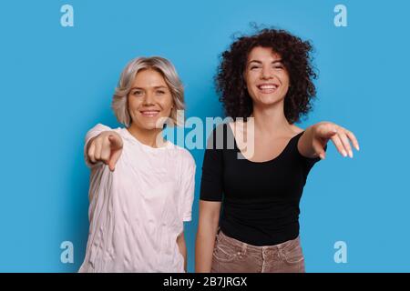 Une femme blonde caucasienne avec son ami aux cheveux bouclés pointe vers l'appareil photo sur un fond bleu Banque D'Images