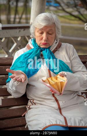 Une femme âgée, triste et habillée avec élégance, est assise sur un banc de parc avec un sac à main vide dans ses mains et une expression confuse sur son visage. Banque D'Images