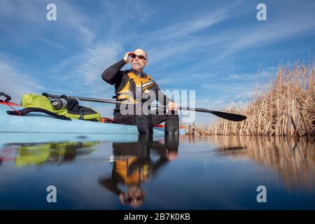 Le paddler masculin senior se détend au soleil de l'après-midi après une séance d'entraînement sur une paddleboard debout, lac dans le Colorado, paysage d'hiver ou de début de printemps, recrea Banque D'Images