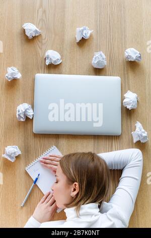 Femme fatiguée freelance dormant et reposant sa tête sur un ordinateur portable sur la table, écrivant sur le bloc-notes, des boules de papier et d'ordinateur portable froissés autour, vue de dessus Banque D'Images