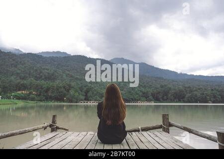 Gros plan image d'une femme assise seule sur un vieux quai en bois près de la rivière avec ciel et fond de montagne Banque D'Images