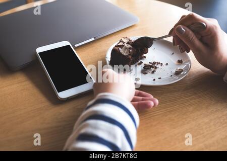 Image Mockup d'un téléphone mobile blanc avec écran noir blanc de bureau à côté d'un ordinateur portable avec une femme mangeant le gâteau de brownie Banque D'Images
