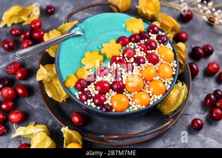 Bol à smoothie aux fruits bleus teint avec de la poudre saine de spiruline naturelle décorée de canneberges, de physalis et de kaki et de grains de quinoa gonflés Banque D'Images