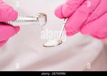 Gros plan les mains du dentiste dans des gants de protection roses avec miroir à bouche métallique et pièce à main dentaire à grande vitesse sur fond flou. Outils médicaux conceps Banque D'Images